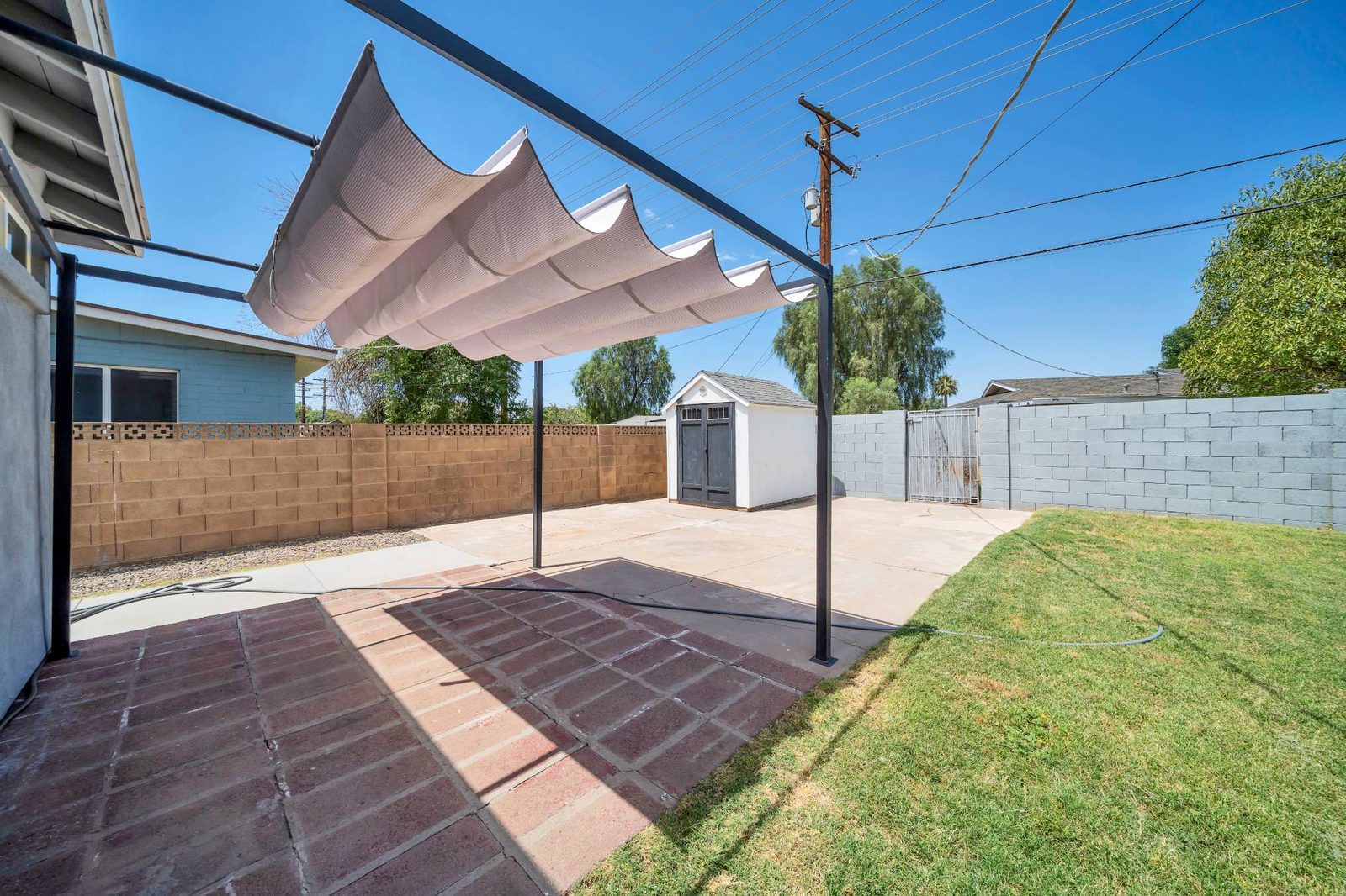 Paved patio under a shade sail, with the storage shed at the far end